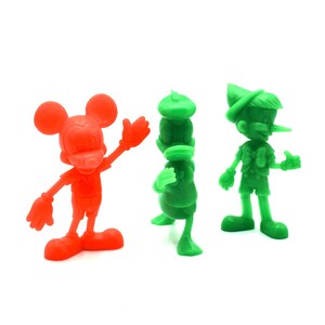May include: Three colorful plastic figurines of cartoon characters: Mickey Mouse in red, Donald Duck in green, and Pinocchio in green. The figures are standing on a white surface.