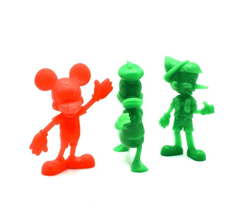 May include: Three colorful plastic figurines of cartoon characters: Mickey Mouse in red, Donald Duck in green, and Pinocchio in green. The figures are standing on a white surface.