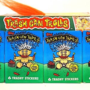 Pu&ograve; includere: Quattro pacchetti di figurine Topps Trash Can Trolls, ognuno con un'illustrazione di un troll dei cartoni animati. I pacchetti sono blu con le parole "Trash Can Trolls" e "6 Trashy Stickers". &Egrave; visibile anche una scatola di carte e capelli arancioni.