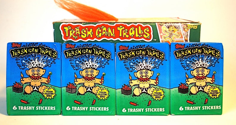 Pu&ograve; includere: Quattro pacchetti di figurine Topps Trash Can Trolls, ognuno con un'illustrazione di un troll dei cartoni animati. I pacchetti sono blu con le parole "Trash Can Trolls" e "6 Trashy Stickers". &Egrave; visibile anche una scatola di carte e capelli arancioni.