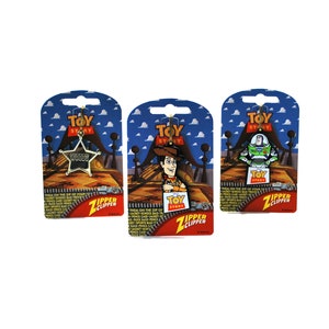 Puede incluir: Tres charms de cremallera de Toy Story en tarjetas de embalaje. Los charms son Woody, Buzz Lightyear y una estrella de sheriff con la palabra "WOODY" en ella. Las tarjetas de embalaje son azules, rojas y amarillas con nubes blancas y el logotipo de Toy Story.