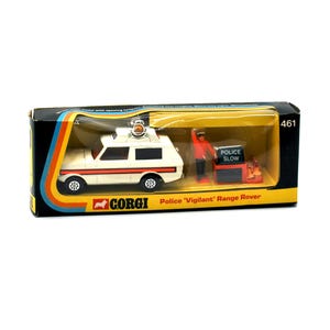 May include: A vintage Corgi Police 'Vigilant' Range Rover toy car set in its original box. The white and red vehicle has a roof light and is accompanied by a police officer figure and a 'Police Slow' sign. The box has a black, yellow, and orange design.