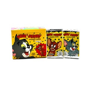 May include: A yellow box of Tom and Jerry trading cards with a picture of Tom on the front. Two packs of Tom and Jerry trading cards are shown in the foreground. The packs are yellow with pictures of Tom and Jerry on the front.