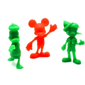 May include: Three colorful plastic figurines of cartoon characters: a green duck-like figure, a red Mickey Mouse, and a green Pinocchio. The figures are standing on a white surface. The Mickey Mouse figure is waving.