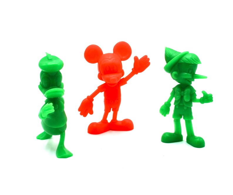 May include: Three colorful plastic figurines of cartoon characters: a green duck-like figure, a red Mickey Mouse, and a green Pinocchio. The figures are standing on a white surface. The Mickey Mouse figure is waving.