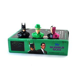 May include: A green box of Topps Batman Forever bubble gum with three plastic figures on top. The figures are Batman, The Riddler, and Two-Face. The box has a picture of Batman, The Riddler, and Two-Face on the front.