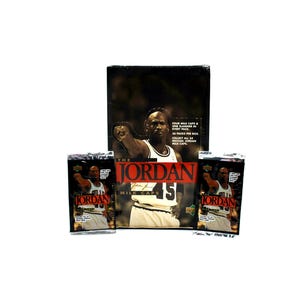 May include: A collection of "The Jordan" milk caps, featuring Michael Jordan. The image shows a box and individual packs with the basketball player's image and the number 45. The box reads "FOUR MILK CAPS & ONE SLAMMER IN EVERY PACK. 36 PACKS PER BOX. COLLECT ALL 54 MICHAEL JORDAN MILK CAPS."