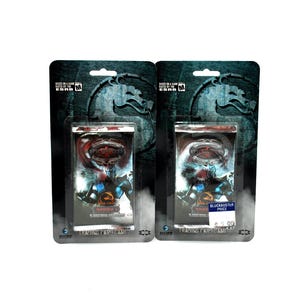 Pode incluir: Dois pacotes lacrados de boosters do jogo de cartas colecionáveis Mortal Kombat. Cada pacote apresenta um cartão azul e preto com um personagem do jogo. Os pacotes estão sobre um fundo branco.
