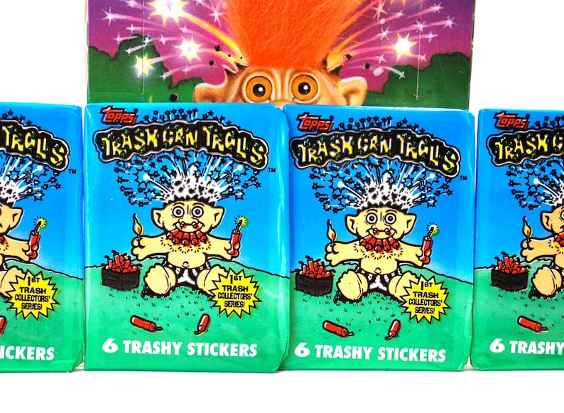 Pu&ograve; includere: Quattro confezioni di adesivi "Trash Can Trolls" di Topps, con un troll dei cartoni animati con i capelli arancioni e un petardo. Le confezioni sono blu e verdi con la scritta "6 Trashy Stickers". Un'immagine pi&ugrave; grande del troll &egrave; visibile sopra.