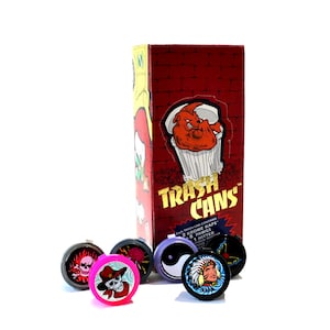 May include: A box of Trash Cans, a brand of smoking accessories. The box is red with a brick pattern and features a cartoon image of a bulldog in a trash can. The box says "Trash Cans" in gold letters. There are six small, colorful, round containers with various designs on them. Each container has a small, round, black lid.