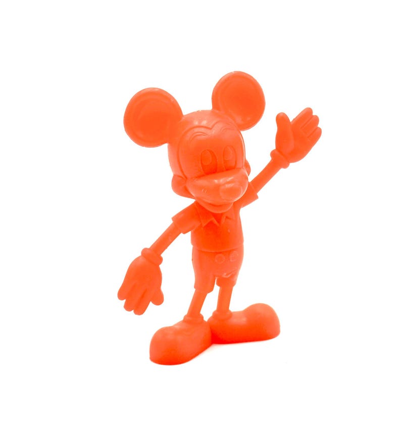 May include: Orange figurine of Mickey Mouse in a classic pose, with one arm raised. The figure has large round ears, a button-up shirt, shorts, and large shoes. The figurine is a solid orange color and stands on a white background.