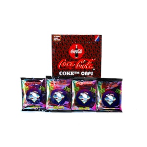 May include: Four sealed packs of Coca-Cola CokeTM Caps trading cards. Each pack contains 5 caps and features a holographic design with a white dolphin on a blue background. The packs are arranged in a row with a box of Coca-Cola CokeTM Caps trading cards behind them. The box is red and black with the Coca-Cola logo and the text "CokeTM Caps".