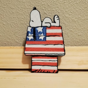 May include: A Snoopy-themed decoration featuring Snoopy lying on top of his doghouse. The doghouse is designed with the American flag, displaying red and white stripes with blue and white stars. The decoration is set against a neutral background.