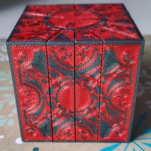 May include: A red and black puzzle box with intricate geometric patterns. The cube-shaped object has a textured surface and appears to be made of a solid material. The design features a central circular element and symmetrical details.