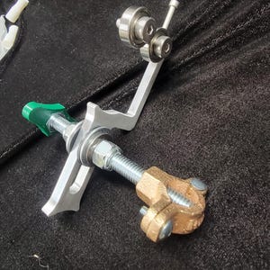 Op de afbeelding: Een zilveren metalen gereedschap met een schroefdraadstang, een groene kunststof handgreep en een bronzen klem. Het gereedschap heeft een gebogen arm met twee zilveren lagers.