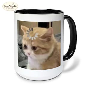 May include: White ceramic mug with a black handle and interior. The mug features a meme of a cat with a loading symbol above its head. The cat is light brown and white with a sad expression. The mug has the shop's logo in the upper left corner.