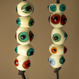 May include: Two vertical strands of black cord featuring handmade glass beads. Each bead is off-white with embedded circular patterns resembling eyes in shades of teal, dark brown, orange, and light blue, creating a multi-coloured, eye-like motif.