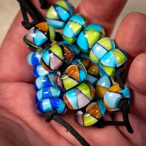 Könnte beinhalten: Eine Sammlung farbenfroher Glasperlen auf schwarzem Kordelband. Die Perlen weisen ein geometrisches Design mit Blau-, Orange-, Gelb- und Rosatönen auf. Die Perlen sind glatt und abgerundet, mit glänzendem Finish.