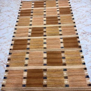 May include: A rectangular area rug featuring a repeating grid pattern of alternating light tan and dark brown squares. The design is accented by vertical stripes of white and dark blue thread that separate the squares, creating a structured, woven appearance.