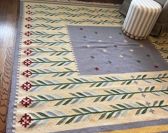 May include: A rectangular area rug with a central gray square and a cream-colored border. The border features a repeating pattern of green and blue leaves. The rug has a fringed edge and is placed on a hardwood floor.