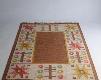 May include: Rectangular woven rug with a brown center and a cream-colored border. The border features repeating geometric patterns in yellow, pink, green, and brown. The rug has a fringed edge and is suitable for a living room or bedroom.