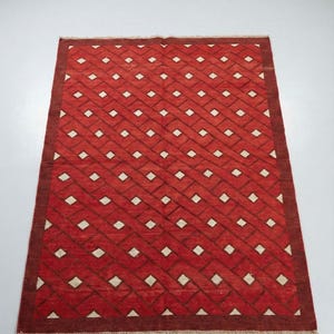 Op de afbeelding: Een rechthoekig rood tapijt met een geometrisch patroon van witte ruiten en donkerrode lijnen. Het tapijt heeft een donkerrode rand en een gefranjerde rand. Het tapijt is waarschijnlijk gemaakt van geweven materiaal.