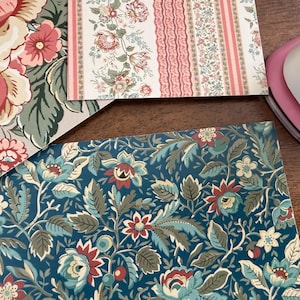 May include: Close-up of patterned paper sheets with floral designs. One sheet features large pink and red roses on a beige background. Another has a blue background with teal and red flowers. A pink paper punch is in the corner.