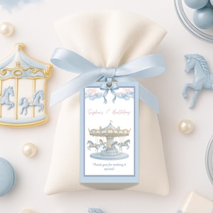 Op de afbeelding: Een witte cadeauzakje vastgebonden met een lichtblauw lint en label. Het label leest "Sophia's 1st Birthday!" met een carrousel illustratie. Decoratieve elementen zijn blauwe carrousel paarden beeldjes, parels en een blauwe macaron.