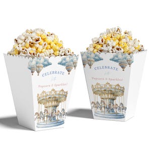 Puede incluir: Dos cajas blancas de palomitas de maíz llenas de palomitas. Las cajas presentan un diseño de carrusel con globos aerostáticos y el texto "¡Celebra con palomitas y chispas!" Las palomitas son una mezcla de granos blancos y amarillos.