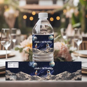 Puede incluir: Botella de agua de plástico transparente con una etiqueta de temática espacial. La etiqueta muestra un cohete, un astronauta, planetas y el texto "Aiden's 7th Birthday" sobre un fondo estrellado. La botella está sobre una mesa.