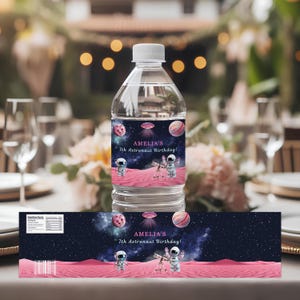 Puede incluir: Una botella de agua de plástico transparente con tapa blanca y etiqueta personalizada. La etiqueta presenta un diseño temático espacial con planetas, un astronauta y el texto "AMELIA'S 7th Astronaut Birthday!"