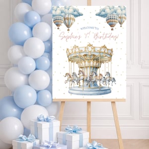 Peut inclure: Un panneau avec une illustration de manège, des montgolfières et le texte "Welcome to Sophie's 3rd Birthday!" est affiché sur un chevalet. Des ballons et des cadeaux emballés bleus et blancs sont en arrière-plan.