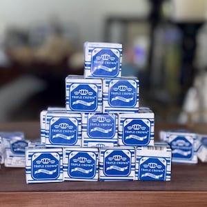 May include: A collection of small, square boxes stacked on a wooden surface. Each box is white with blue accents and features the text "TRIPLE CROWN ORIGINAL" in a stylized design. The boxes are arranged in a pyramid shape, with some scattered around.