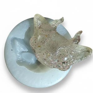 May include: A clear, glitter-infused resin wolf head sits atop a light blue, circular mold. The wolf head is detailed with visible ears and facial features. The mold is a light blue color, and the background is white.