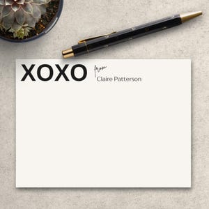 May include: A white stationery card with the text "XOXO from Claire Patterson" in black. A black pen with gold accents rests on the card. A small potted succulent is in the upper left corner.