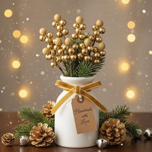 Peut inclure: Un vase blanc avec un ruban doré et une étiquette « Handmade with Love » contient des tiges de baies dorées et de la verdure. L'arrangement est entouré de pommes de pin, de cloches et de lumières, créant une ambiance festive.