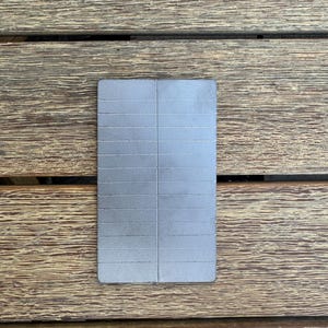 Può includere: Un oggetto rettangolare color argento con una serie di linee orizzontali, possibilmente un magnete o una piastra metallica. L'oggetto è appoggiato su una superficie di legno con un motivo a venature naturali.