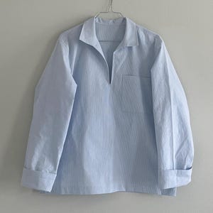 May include: A light blue long-sleeve shirt with a subtle vertical stripe pattern, featuring a pointed collar, a deep V-neckline, and a single chest pocket on the left side. The shirt has cuffed sleeves and is displayed on a metal wire hanger.