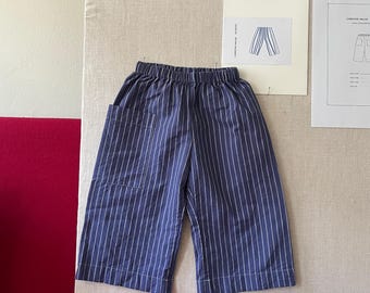 May include: A pair of navy blue cropped pants featuring a vertical white pinstripe pattern, an elasticized waistband, and a large patch pocket on the right side. The pants have a wide-leg silhouette and a straight hemline, constructed from fabric.