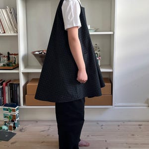 Puede incluir: Un vestido con delantal a cuadros negros y grises con una camiseta blanca debajo. El vestido tiene un bolsillo grande en la parte delantera y se combina con pantalones negros. Adecuado para ropa de niños.