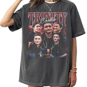 Könnte beinhalten: Ein anthrazitfarbenes Kurzarm-T-Shirt mit einem Grafikdruck, der den Text "TRINITY" in großen roten Buchstaben und "Santos" in weißer Schreibschrift zeigt. Das Design enthält mehrere Bilder einer Person in medizinischer Arbeitskleidung.