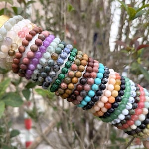 Op de afbeelding: Een verzameling kralenarmbanden in verschillende kleuren, waaronder roze, paars, groen en blauw. De armbanden zijn gemaakt van kleine, ronde kralen en zijn op elkaar gestapeld. De achtergrond is wazig, met groen gebladerte zichtbaar.