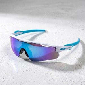 May include: Performance sunglasses featuring a white frame with bright blue temple tips and a single-piece wrap-around lens. The lens has a reflective purple and blue gradient finish with the word Prizm etched into the lower right corner.
