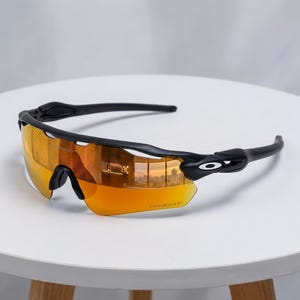 May include: Performance sunglasses featuring a matte black frame and a single-piece wrap-around lens with a reflective orange tint. The lens includes the Prizm logo etched in the corner, and the temple arms display the white Oakley O logo.
