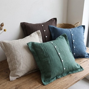 May include: Four square throw pillows in shades of beige, brown, blue, and green. Each pillow has a buttoned closure down the center. The pillows are made of a textured fabric and rest on a wooden surface.