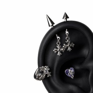 May include: A black ear model displaying silver-toned jewelry including an industrial barbell with two spikes and two dangling cross charms, a silver cross-shaped ear cuff with a clear stone, and a purple heart-shaped stud with a silver cross overlay.