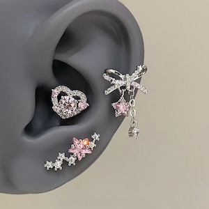 May include: A collection of silver-toned earrings with pink gemstones. The earrings include a heart-shaped stud, a bow-shaped ear cuff with dangling stars, and a star-shaped climber earring. The jewelry is set against a gray background.