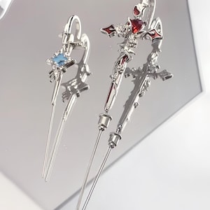 May include: Two silver hair sticks with decorative tops. One features a blue gemstone, the other a red gemstone. Both have a cross design with intricate details and long, thin shafts.