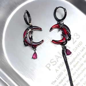 May include: A pair of dark metal earrings featuring a crescent moon design with red enamel and a dangling teardrop-shaped gem. The earrings are on a silver tray.