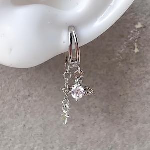 May include: A silver-toned hoop earring featuring two dangling charms. One charm is a circular orb shape with a central clear stone, and the other is a delicate chain ending in a small, four-pointed star. The metal has a polished finish.